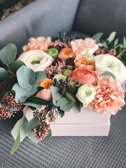 A floral arrangement featuring a variety of fresh flowers in soft pastel shades, including blush pink and cream ranunculus, peach and apricot carnations, and green buds, artistically arranged in a whi