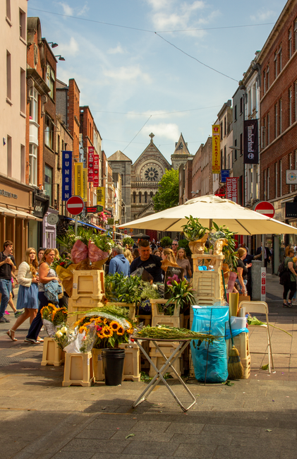 A vibrant outdoor flower market on Guildford High Street, featuring various floral arrangements and bouquets displayed on wooden crates and tables under a large cream parasol. The flowers include brig