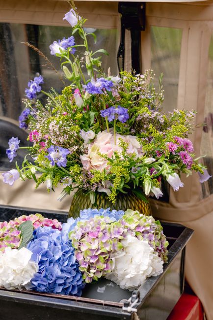 A vibrant floral arrangement featuring a mix of fresh flowers including light pink roses, blue hydrangeas, purple and white lisianthus, and delicate green foliage, arranged in a brown glass vase. The