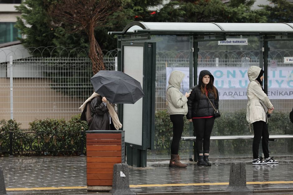 A woman holding a grey umbrella stands on a rainy sidewalk near a bus shelter, which contains three women dressed in hooded jackets and coats waiting in front of a glass wall. The bus shelter features